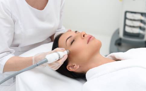 Beautiful girl is receiving anti-aging rejuvenation procedure at spa salon. She is lying with pleasure. Beautician is lifting laser near her cheek in fabrx